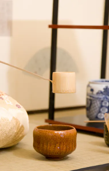 Tea Ceremony A person in a traditional kimono prepares tea, holding a bamboo ladle over a ceramic bowl, with tea ceremony utensils arranged neatly on a tatami mat.