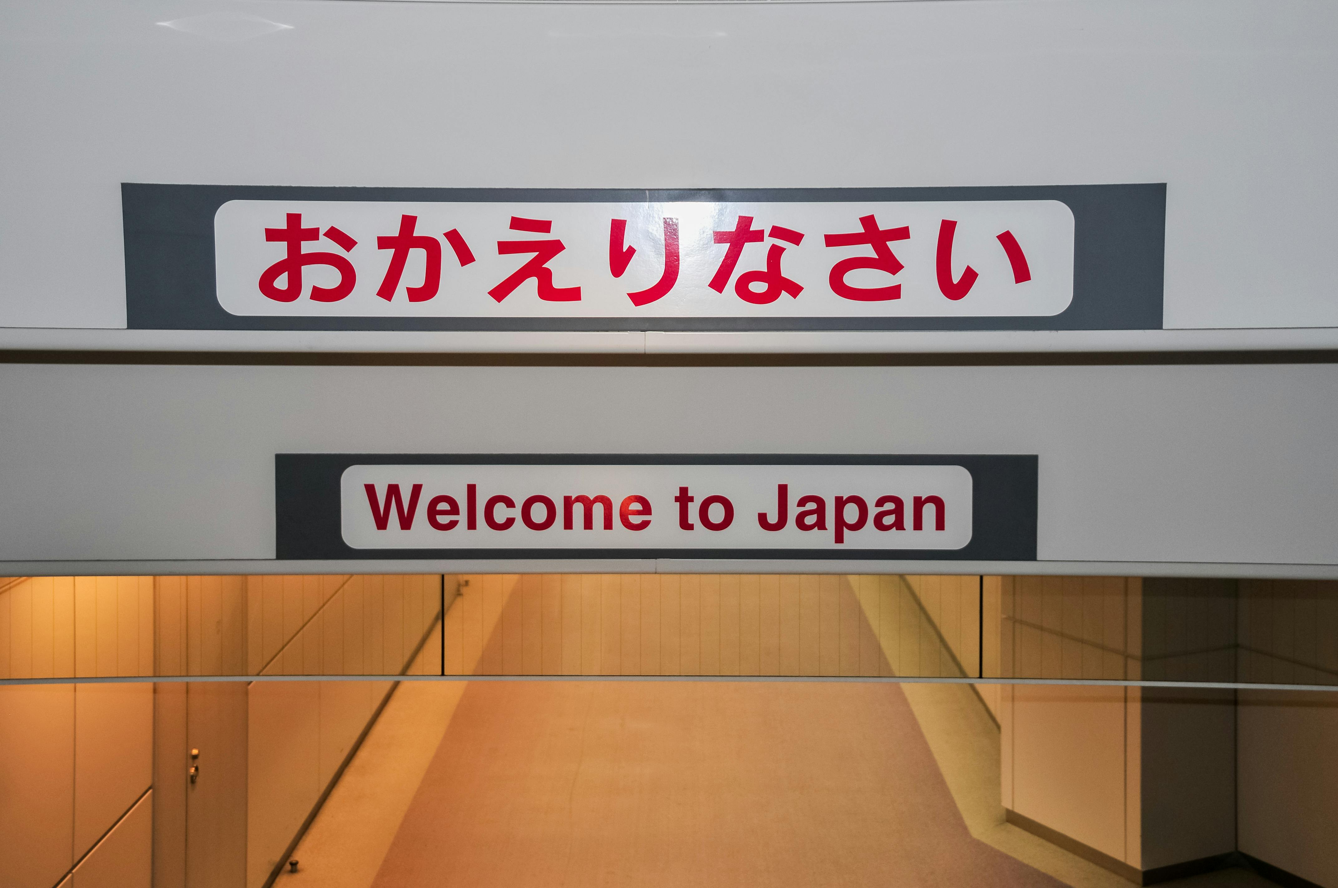 A sign above an airport hallway displays “おかえりなさい” in Japanese and “Welcome to Japan” in English, greeting arrivals. The hallway below is empty and softly lit.