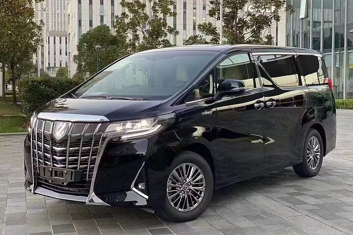 A black luxury minivan with tinted windows and chrome accents is parked on a paved area in front of modern office buildings and trees.