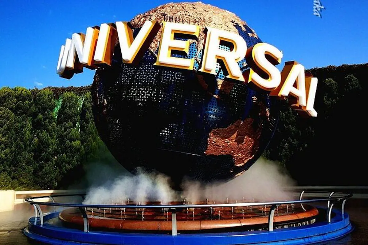 The image shows a large globe with the word "UNIVERSAL" in bold letters wrapped around it. The globe is surrounded by mist and set against a backdrop of trees and a bright blue sky.