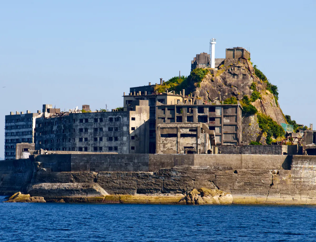 Hashima A deserted, concrete island with abandoned buildings and a lighthouse on a hill, surrounded by blue ocean under a clear sky.
