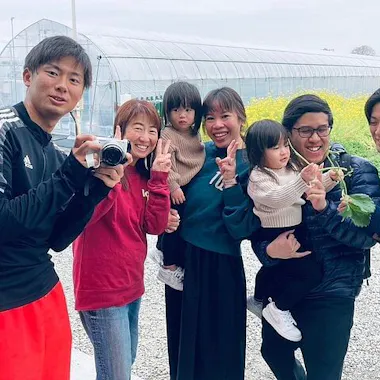 Izumisano Strawberry Picking A group of seven people, including two young children, pose and smile together outdoors near a greenhouse. One person holds a camera, and another holds a green leafy vegetable. Everyone appears happy.
