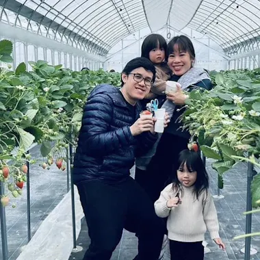 Izumisano Strawberry Picking A family of four, including two young children, poses smiling among rows of strawberry plants in a greenhouse, holding cups, likely for picking or tasting strawberries.