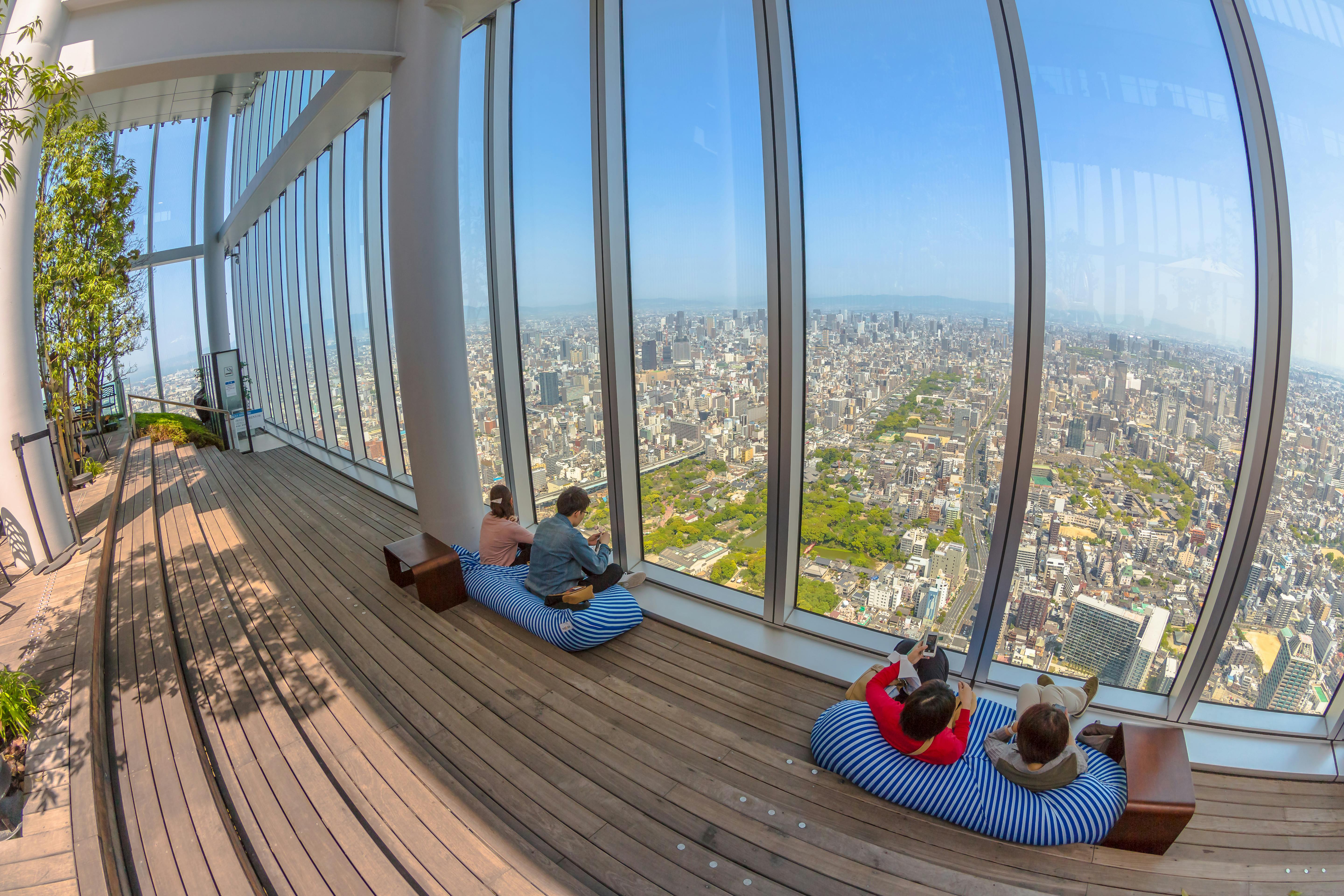 People relax on cushioned seats by large windows in a tall observation deck, enjoying a panoramic city view on a sunny day. The scene has wooden floors, greenery, and a vast urban landscape below.