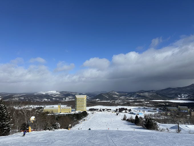 Ski slope with people skiing, leading down to yellow buildings and a snowy resort area, surrounded by trees, mountains, and a partly cloudy blue sky.