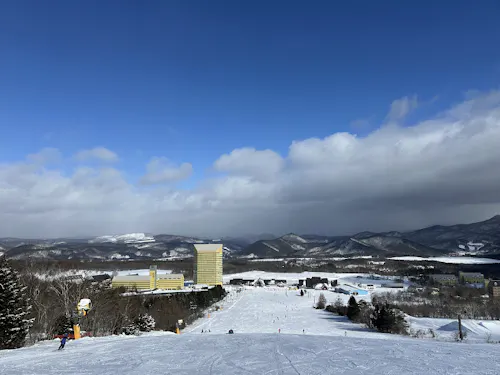 Appi Kogen Ski Resort Ski slope with people skiing, leading down to yellow buildings and a snowy resort area, surrounded by trees, mountains, and a partly cloudy blue sky.