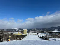 Ski slope with people skiing, leading down to yellow buildings and a snowy resort area, surrounded by trees, mountains, and a partly cloudy blue sky.