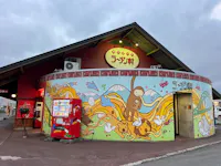 A round red ramen shop with a colorful mural of animals, noodles, Mount Fuji, and waves. A vending machine stands by the entrance under a sign with Japanese text. The sky is cloudy.
