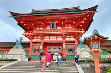 People in colorful traditional clothing walk up the steps toward a large red Shinto shrine gate, with fox statues on either side, under a cloudy sky.