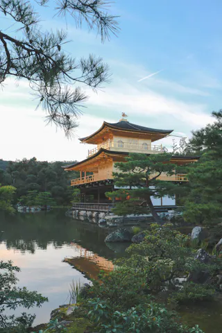 A golden pavilion stands beside a tranquil pond, surrounded by lush greenery and pine trees, under a clear sky.