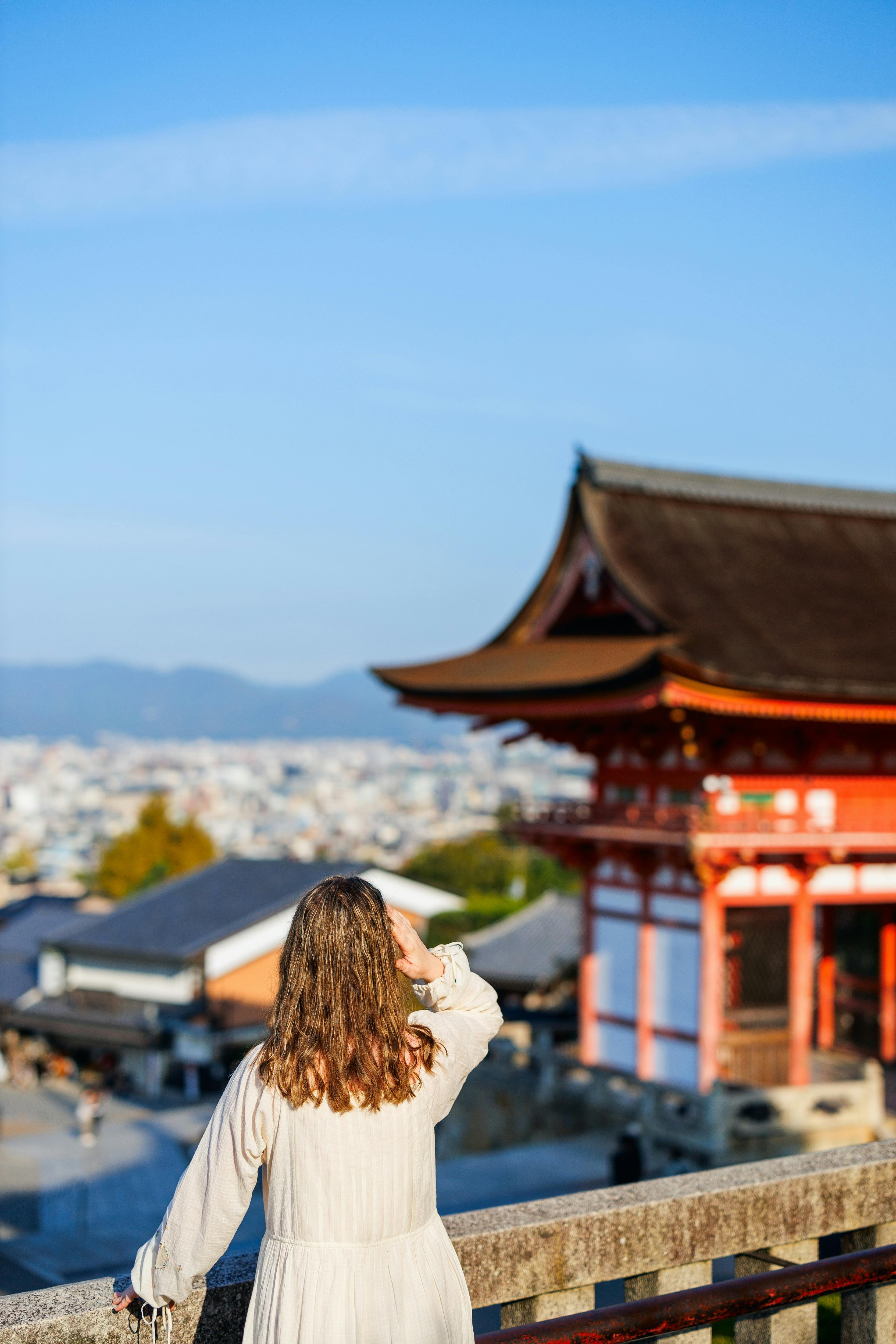 A woman in a white dress stands with her back to the camera, looking at a traditional Japanese temple building with a curved roof, under a clear blue sky. The cityscape and distant mountains are visible in the background.