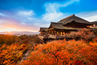 A large traditional Japanese temple overlooks vibrant autumn trees with orange and red leaves at sunset, under a dramatic blue sky with clouds.