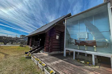 A log cabin stands beside a large glass display case containing an oversized pocket knife. Flower boxes line a wooden walkway under a blue, lightly clouded sky.