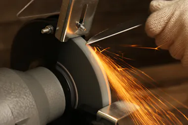 A gloved hand sharpens a knife blade on a spinning grinding wheel, sending bright orange sparks flying off the edge of the blade.