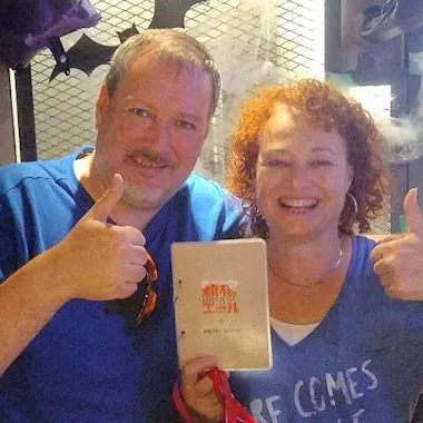 Ramen Making Two smiling people stand close together, giving thumbs up. The person on the right holds a white menu with "Sunset" written on it. Both are wearing blue shirts and are in a casual indoor setting with a metallic background.