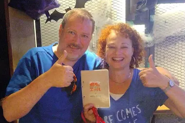 Two smiling people stand close together, giving thumbs up. The person on the right holds a white menu with "Sunset" written on it. Both are wearing blue shirts and are in a casual indoor setting with a metallic background.