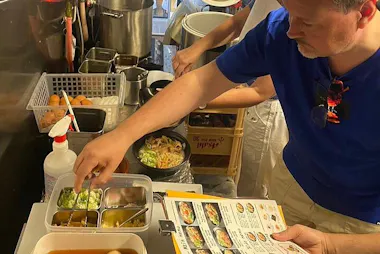 A man in a blue shirt points at a container of toppings at a food counter, holding a menu in one hand. Behind the counter, bowls of soup and cooking pots are visible, along with various kitchen items.