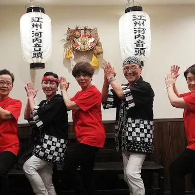 Bon Odori Japanese Folk Dance Five adults pose in a row, dressed in coordinated red and black festival attire, smiling with hands raised. They stand inside a room with Japanese lanterns and a festive wall decoration behind them.