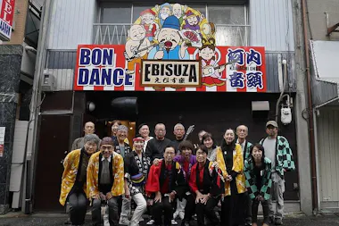 A group of people in colorful traditional clothing pose together outside a building with a sign that reads “EBISUZA,” decorated with cheerful cartoon figures and "BON DANCE" above the entrance.