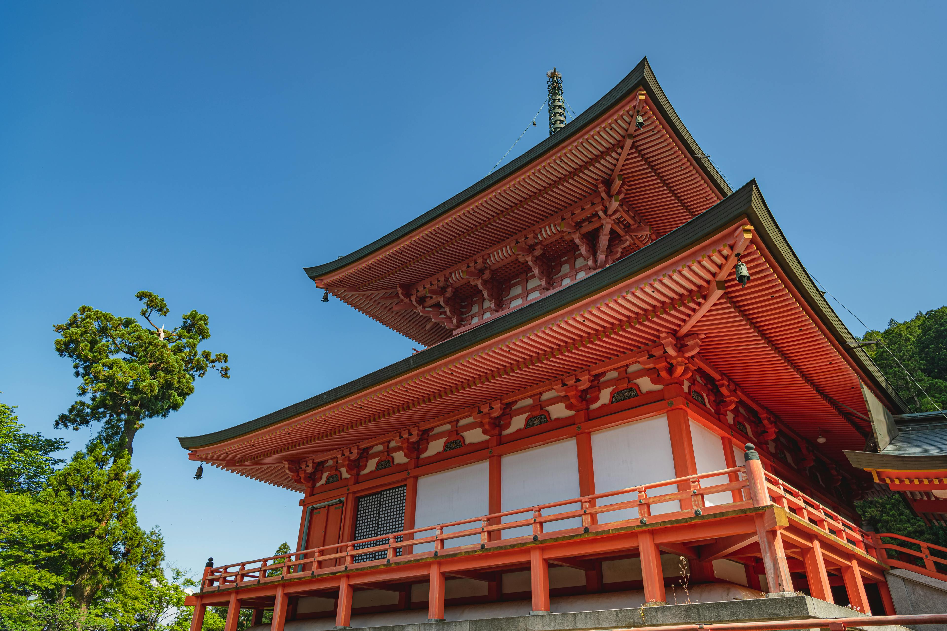 A traditional Japanese pagoda with bright red wooden beams and intricate roof details stands under a clear blue sky, surrounded by lush green trees.