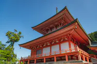 A traditional Japanese pagoda with bright red wooden beams and intricate roof details stands under a clear blue sky, surrounded by lush green trees.