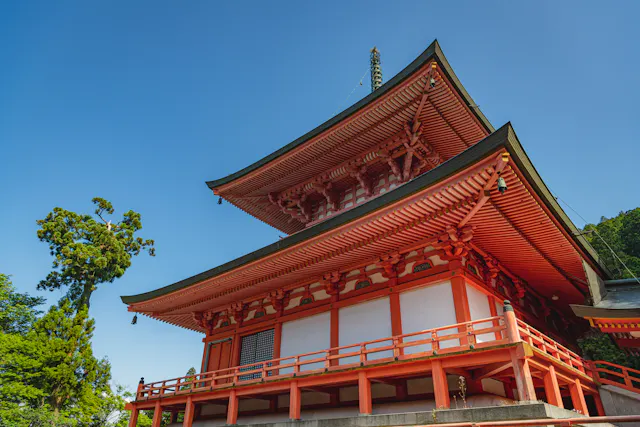 A traditional Japanese pagoda with bright red wooden beams and intricate roof details stands under a clear blue sky, surrounded by lush green trees.