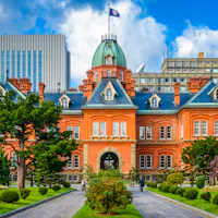 Former Hokkaido Government Building Historic red-brick building with a green dome, surrounded by manicured gardens and trees, set against a backdrop of modern office buildings under a blue sky with clouds.