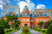 Historic red-brick building with a green dome, surrounded by manicured gardens and trees, set against a backdrop of modern office buildings under a blue sky with clouds.