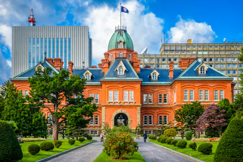 Historic red-brick building with a green dome, surrounded by manicured gardens and trees, set against a backdrop of modern office buildings under a blue sky with clouds.
