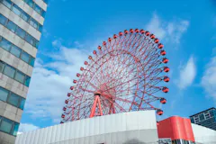 A large red Ferris wheel stands beside a modern building under a bright blue sky with scattered clouds.