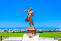 Bronze statue of a man in a long coat pointing into the distance, standing on a pedestal inscribed with "Boys Be Ambitious," with a cityscape and green fields under a clear blue sky in the background.