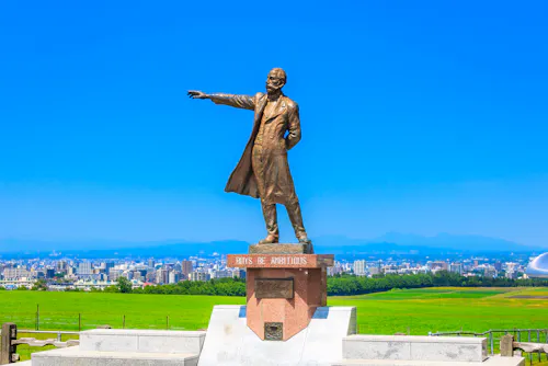 Bronze statue of a man in a long coat pointing into the distance, standing on a pedestal inscribed with "Boys Be Ambitious," with a cityscape and green fields under a clear blue sky in the background.
