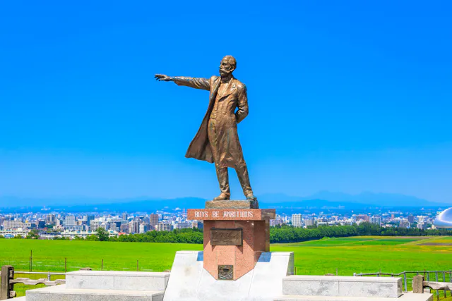 Bronze statue of a man in a long coat pointing into the distance, standing on a pedestal inscribed with "Boys Be Ambitious," with a cityscape and green fields under a clear blue sky in the background.
