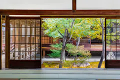 View from inside a traditional Japanese room with tatami flooring, looking out through wooden sliding doors onto a serene garden with green and yellow-leaved trees and a wooden building in the background.