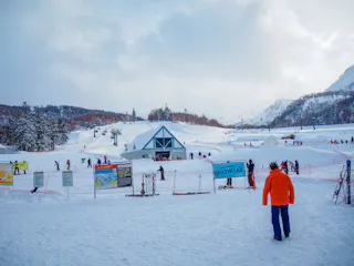 A snowy ski resort with people skiing and walking. Signs and ski equipment are in the foreground, with a blue-roofed building at the center and snow-covered mountains in the background.