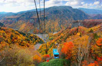 A blue cable car ascends over a forested mountain landscape with vibrant autumn foliage in shades of orange, yellow, and red, under a partly cloudy sky.