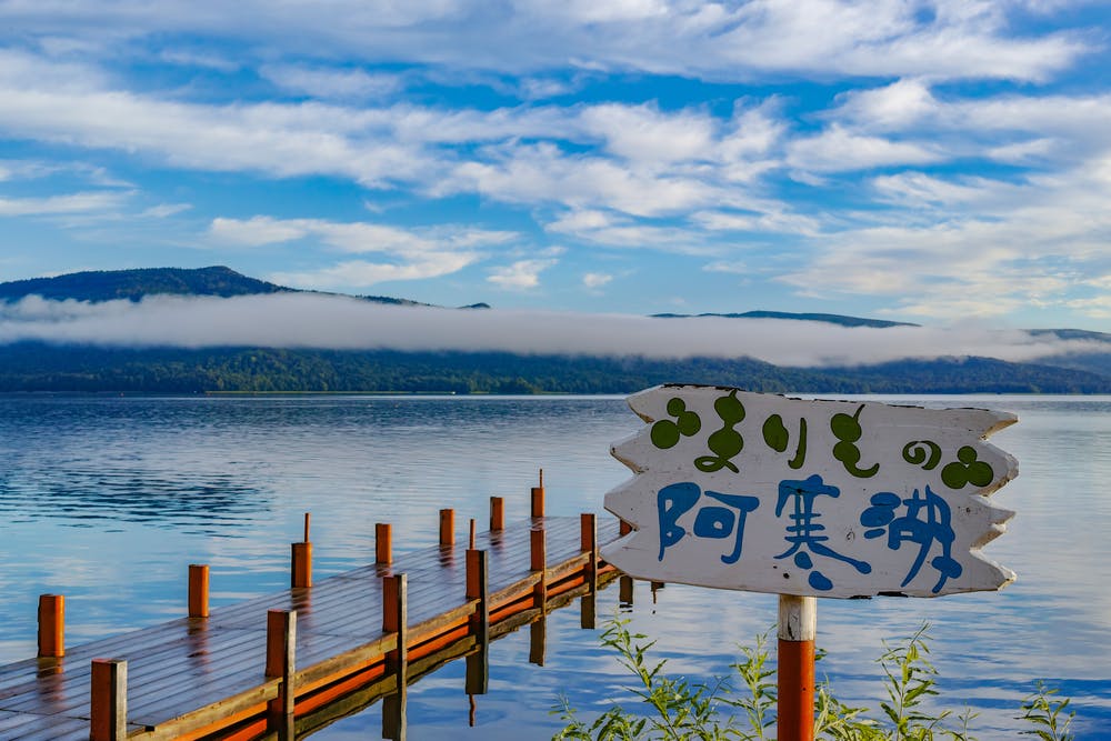 A wooden pier extends into a calm lake with mountains and low-hanging clouds in the background. A white sign with Japanese writing is in the foreground, surrounded by green plants.