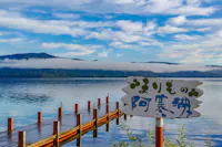 A wooden pier extends into a calm lake with mountains and low-hanging clouds in the background. A white sign with Japanese writing is in the foreground, surrounded by green plants.