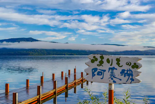 A wooden pier extends into a calm lake with mountains and low-hanging clouds in the background. A white sign with Japanese writing is in the foreground, surrounded by green plants.
