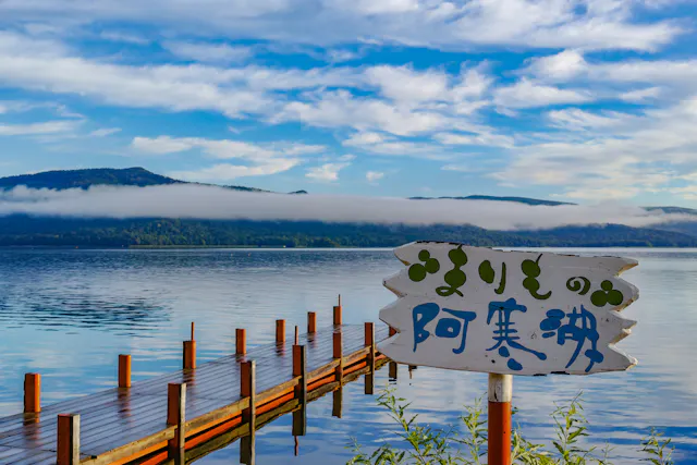A wooden pier extends into a calm lake with mountains and low-hanging clouds in the background. A white sign with Japanese writing is in the foreground, surrounded by green plants.