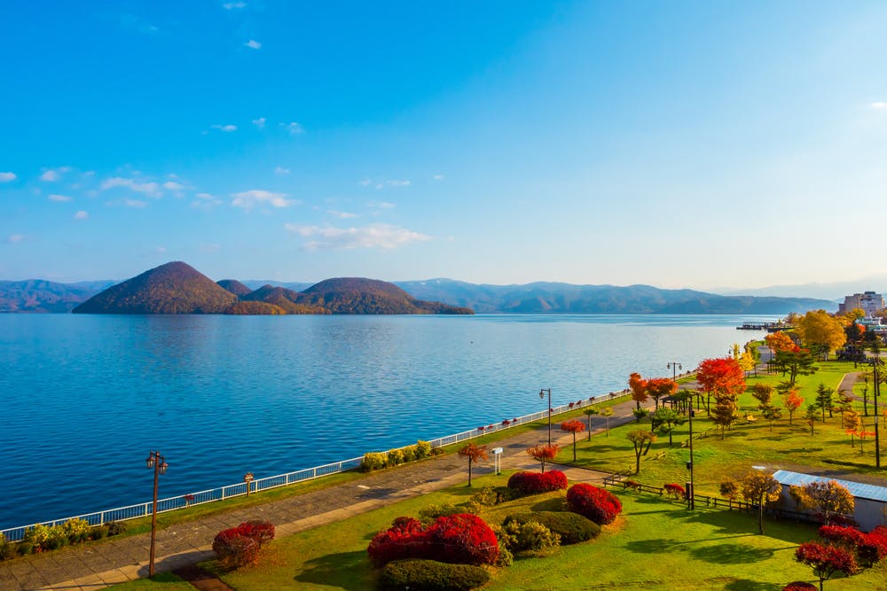 A lakeside park with autumn trees, red and yellow foliage, a walking path, and distant green hills under a clear blue sky with scattered clouds.
