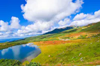 A vibrant landscape featuring a clear blue sky with fluffy clouds, a small lake reflecting the sky, and green hills covered with patches of red and yellow foliage, with distant mountains in the background.