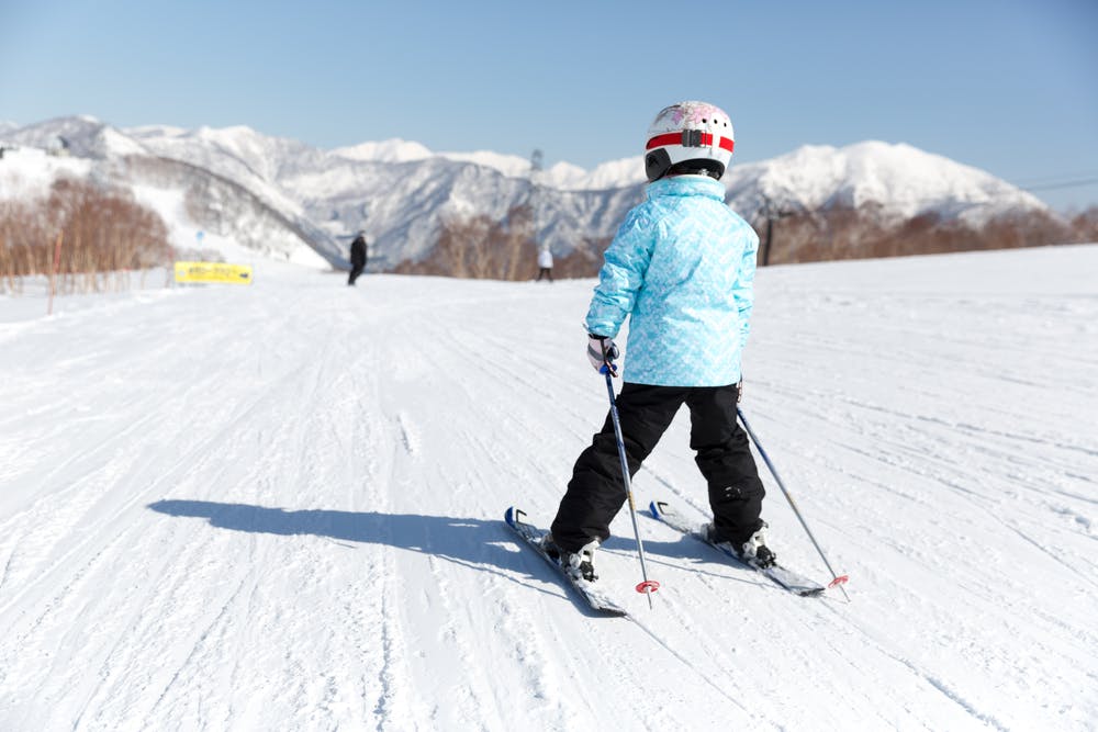 A child wearing a blue jacket, black pants, and a helmet skies on a snowy slope with mountains in the background under a clear blue sky.