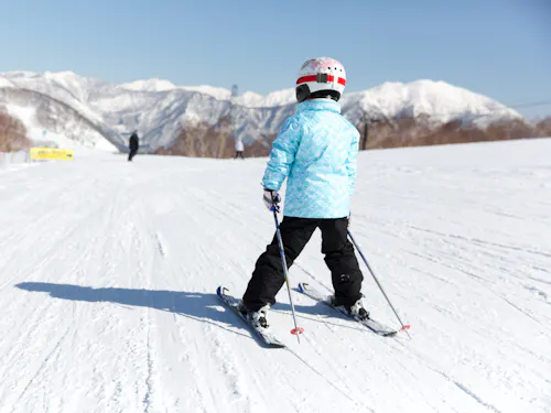 Naeba Ski Resort A child wearing a blue jacket, black pants, and a helmet skies on a snowy slope with mountains in the background under a clear blue sky.