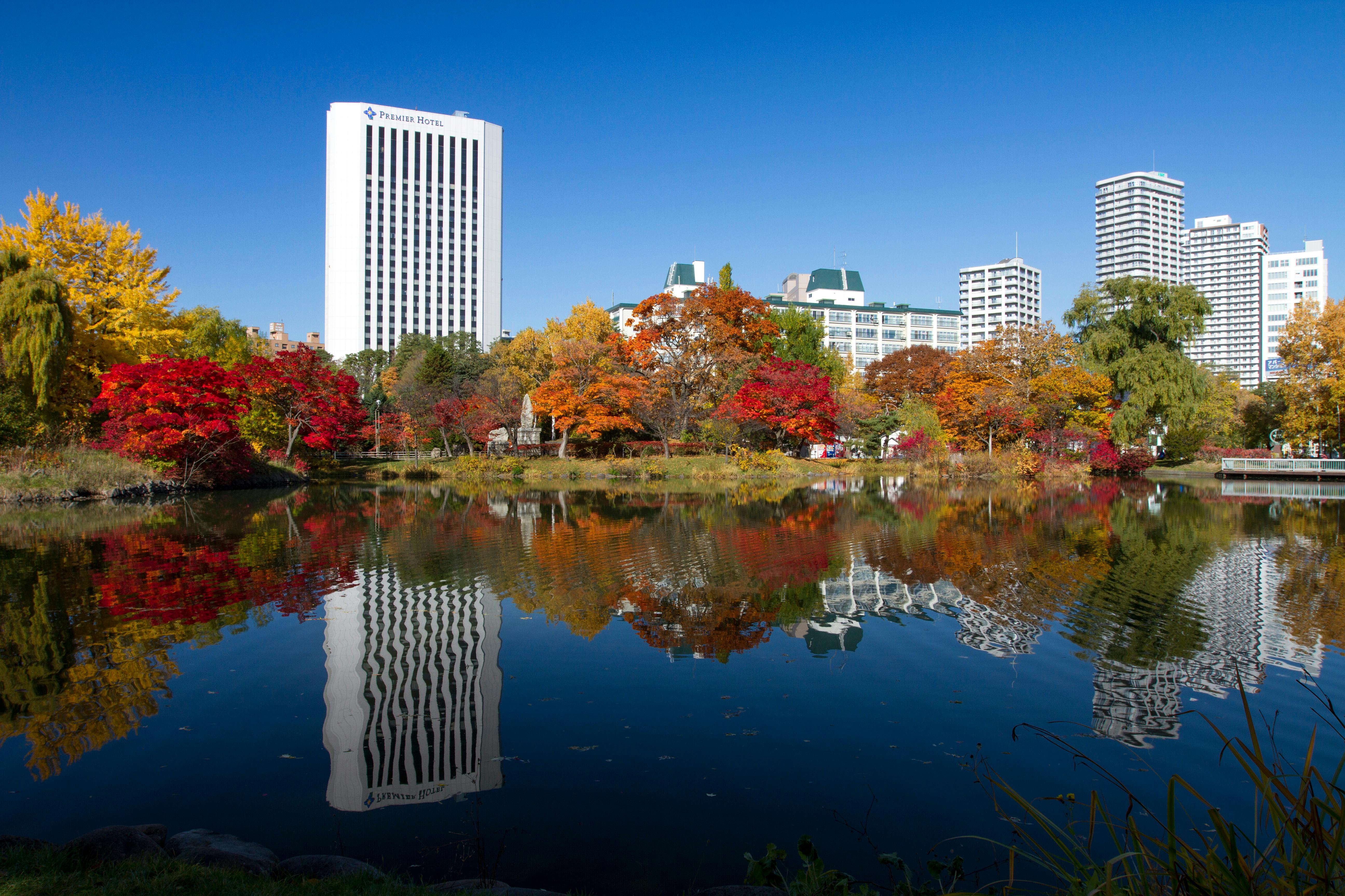 A city park with colorful autumn trees reflected in a calm pond, tall white buildings in the background, and a clear blue sky above.