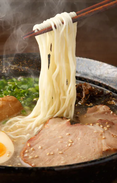 Ramen A steaming bowl of ramen with sliced pork, soft-boiled egg, green onions, bamboo shoots, and sesame seeds, with chopsticks lifting noodles above the broth.