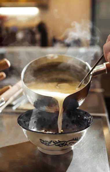 Ramen A hand pours steaming hot soup or broth from a saucepan into a decorative bowl on a kitchen counter, with visible steam rising from the liquid.