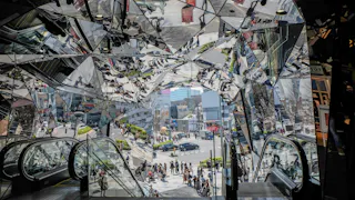 A view from inside a building with mirrored, angular walls reflecting people, escalators, and the street outside, creating a kaleidoscopic effect over an urban scene with pedestrians and city buildings.