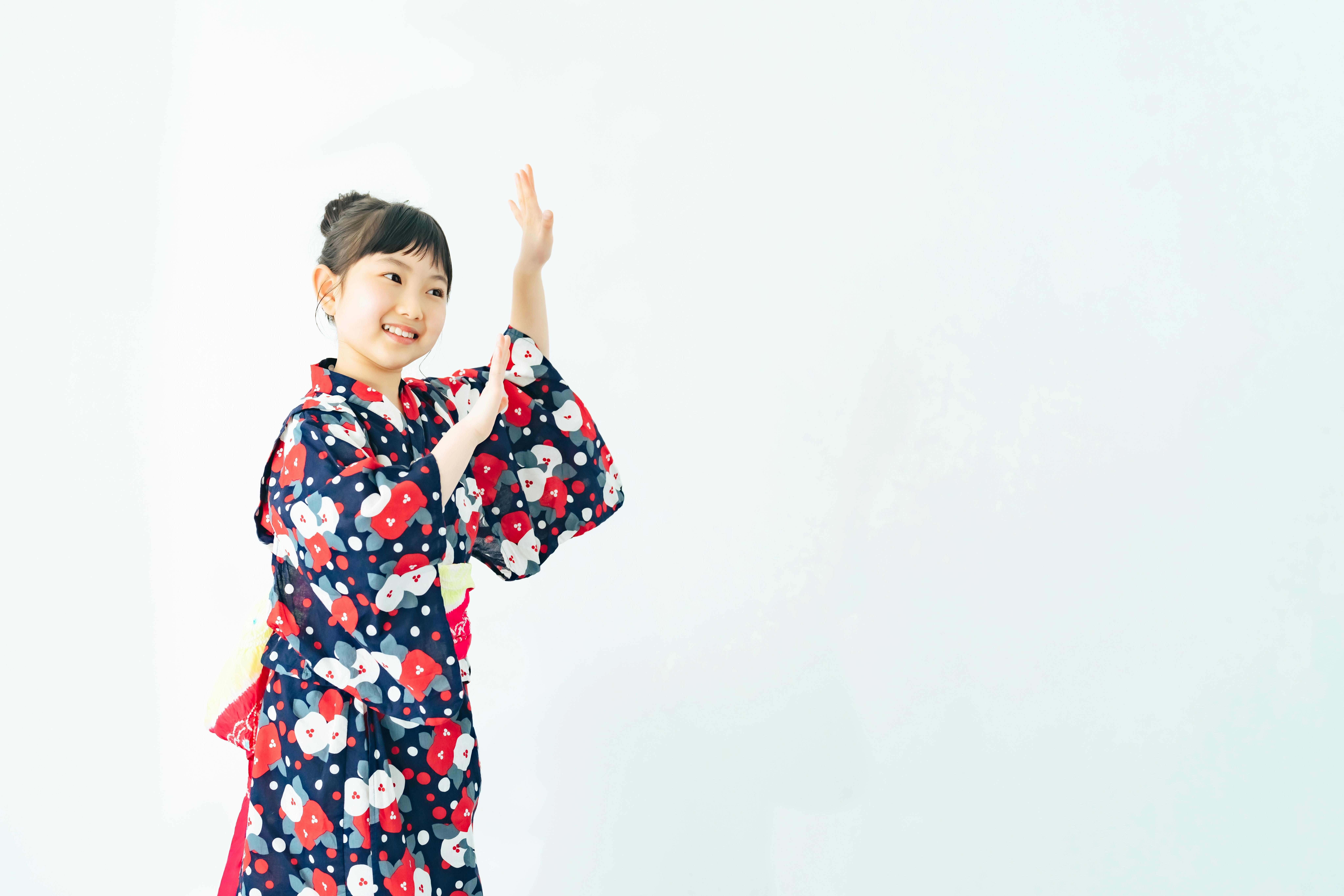 A young girl wearing a colorful yukata with red, white, and blue patterns smiles and raises her hands as if dancing, standing against a plain white background.