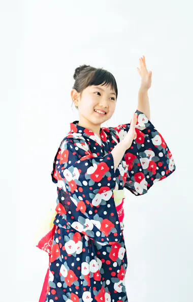 Bon Odori A young girl wearing a colorful yukata with red, white, and blue patterns smiles and raises her hands as if dancing, standing against a plain white background.